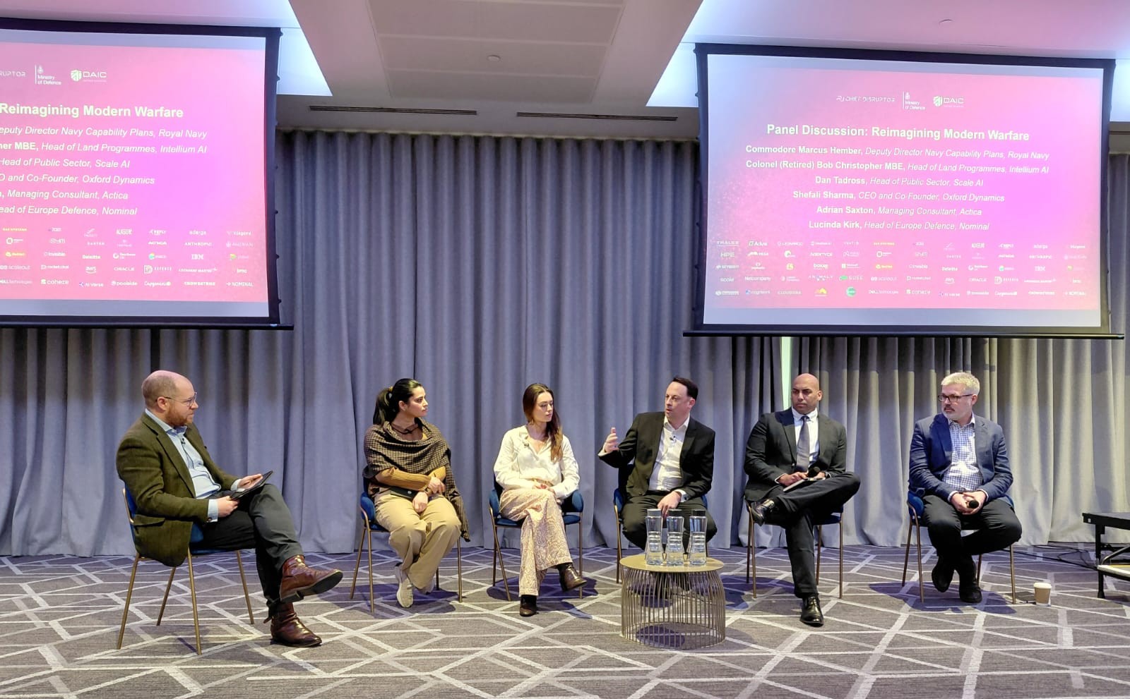 Six people seated in a row on a low stage in front of two screens showing the panel title, Reimagining Modern Warfare, and the panellists' names. Adrian Saxton, third from the right, is speaking, one hand raised.